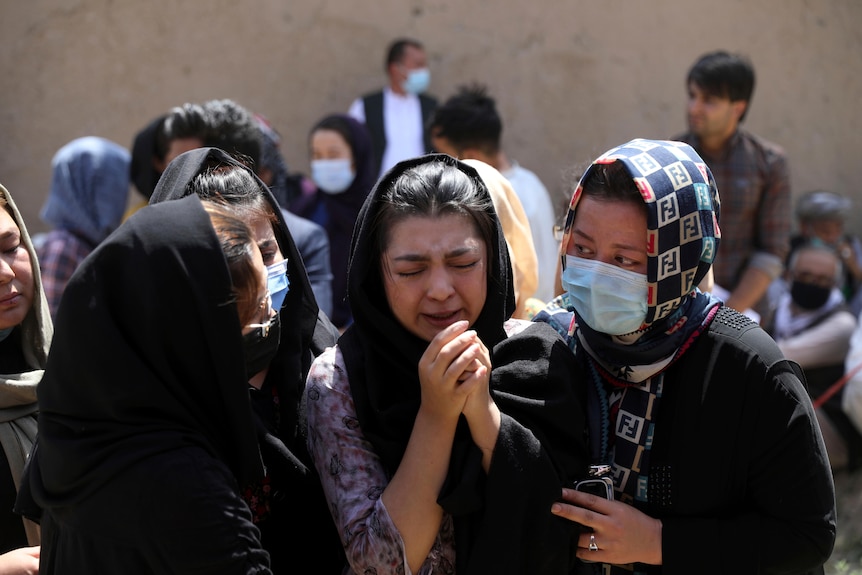 A crying young woman in a headscarf is comforted by three other women, who are wearing headscarves and coronavirus face masks.