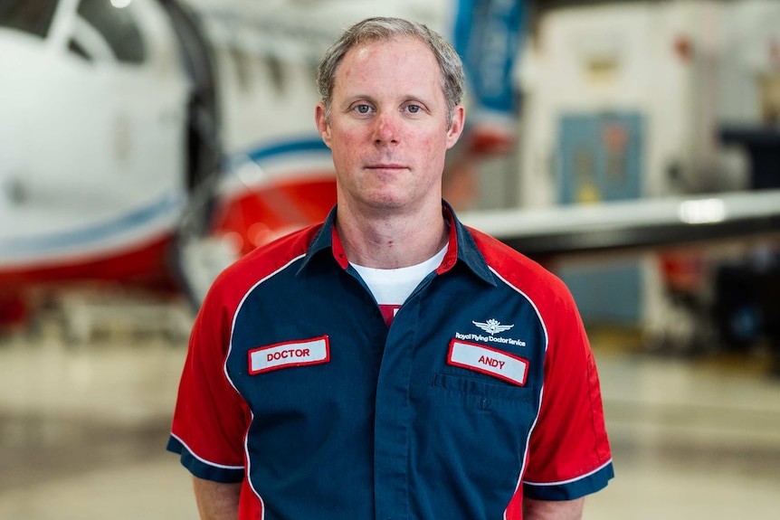 A mid shot of Royal Flying Doctor Service doctor Andy Hooper wearing his red and blue uniform standing in front of a plane.