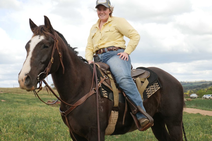 A woman wearing a yellow shirt, blue jeans and cap sitting on a brown horse.