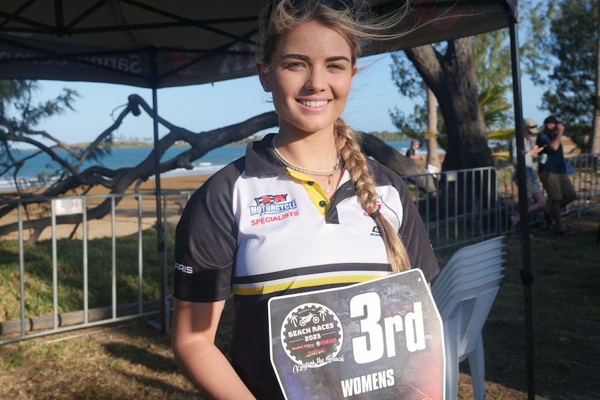 A young woman stands smiling with the beach in the background holding a 3rd place sign. 