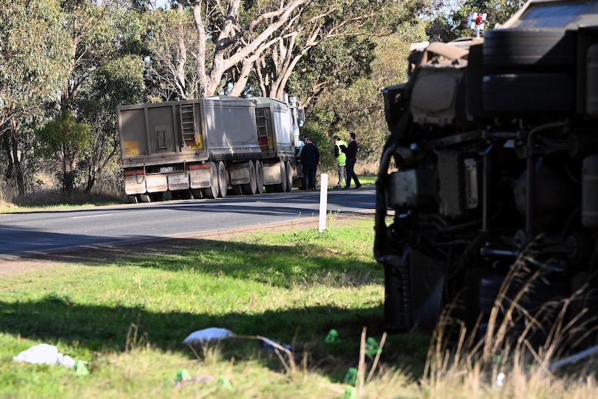 A truck parked on the side of the road in focus, with a truck overturned on its side in the foreground.