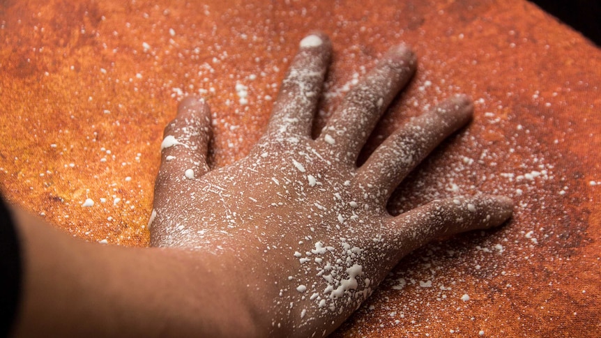 A hand flecked with paint rests on an orange coloured canvas.