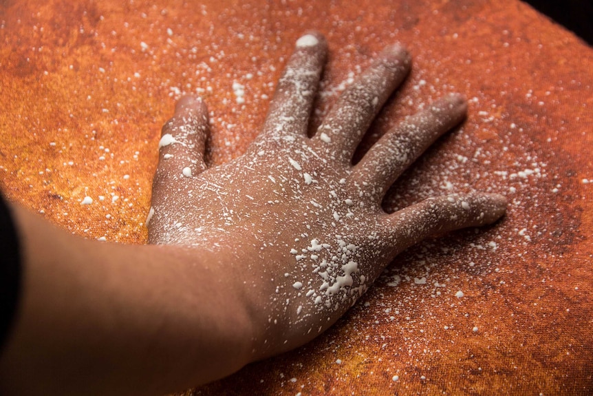 A hand flecked with paint rests on an orange coloured canvas.