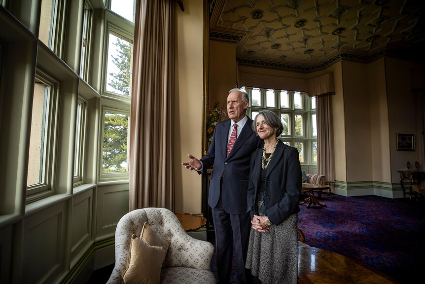 Two middle aged people stand in an ornate room and look out a window