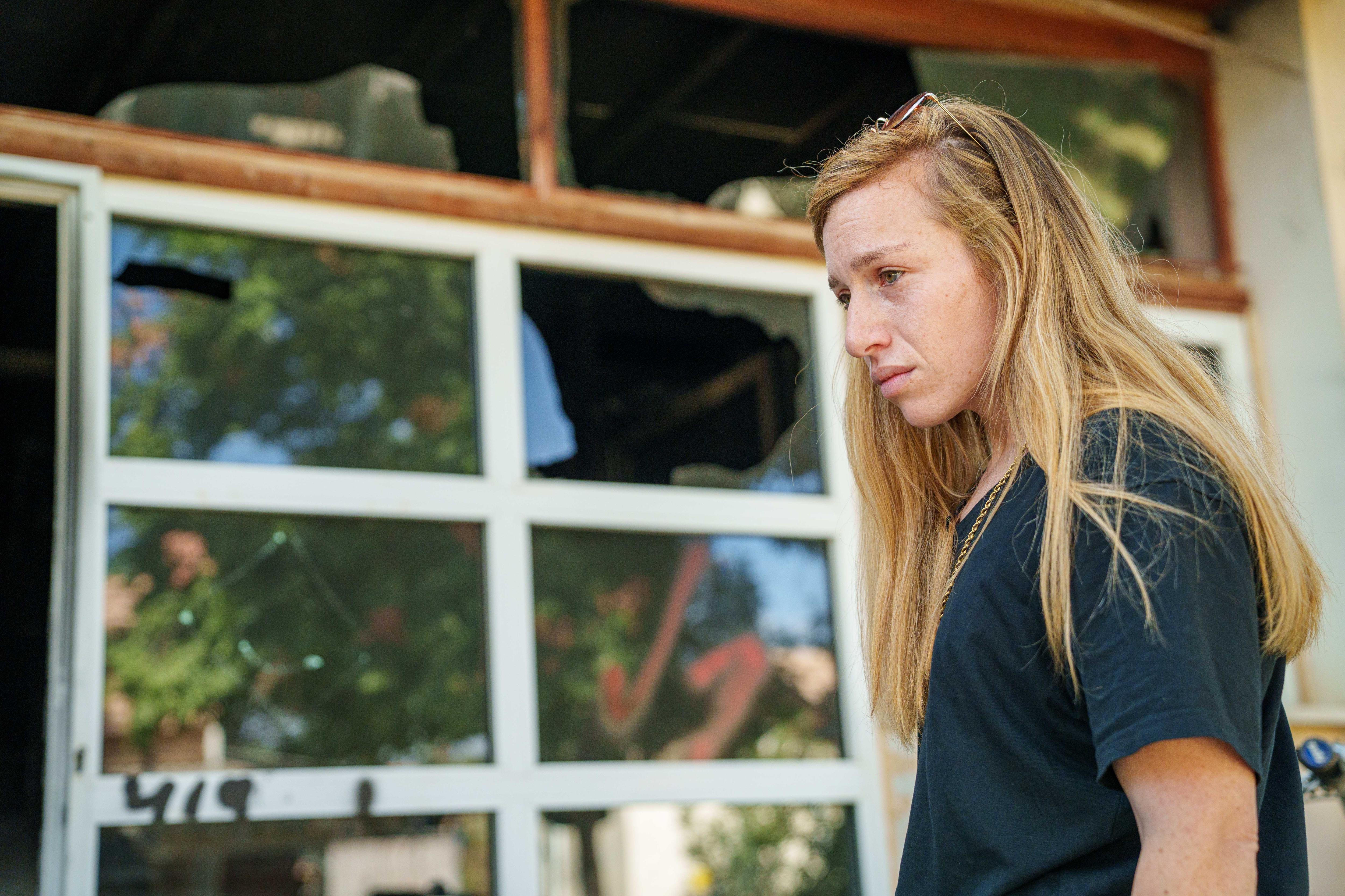A blonde woman stands next to a smashed window 