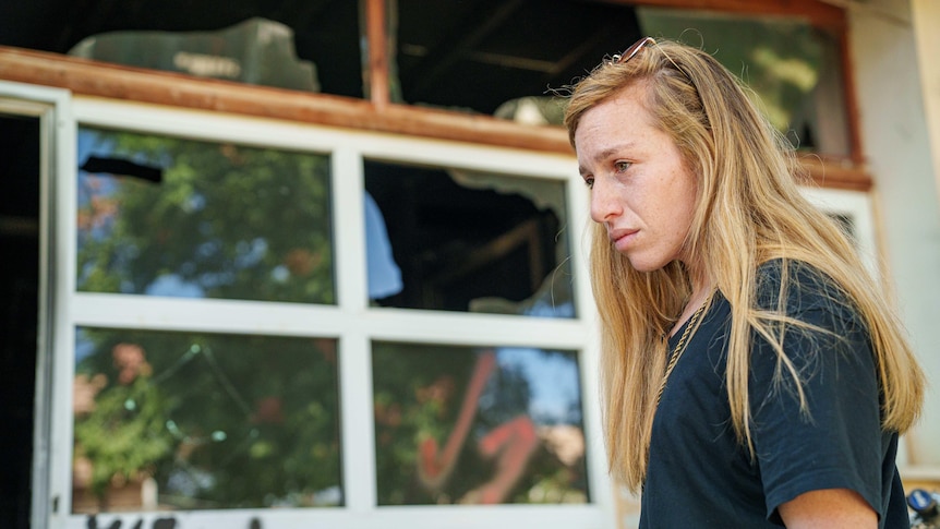 A blonde woman stands next to a smashed window 
