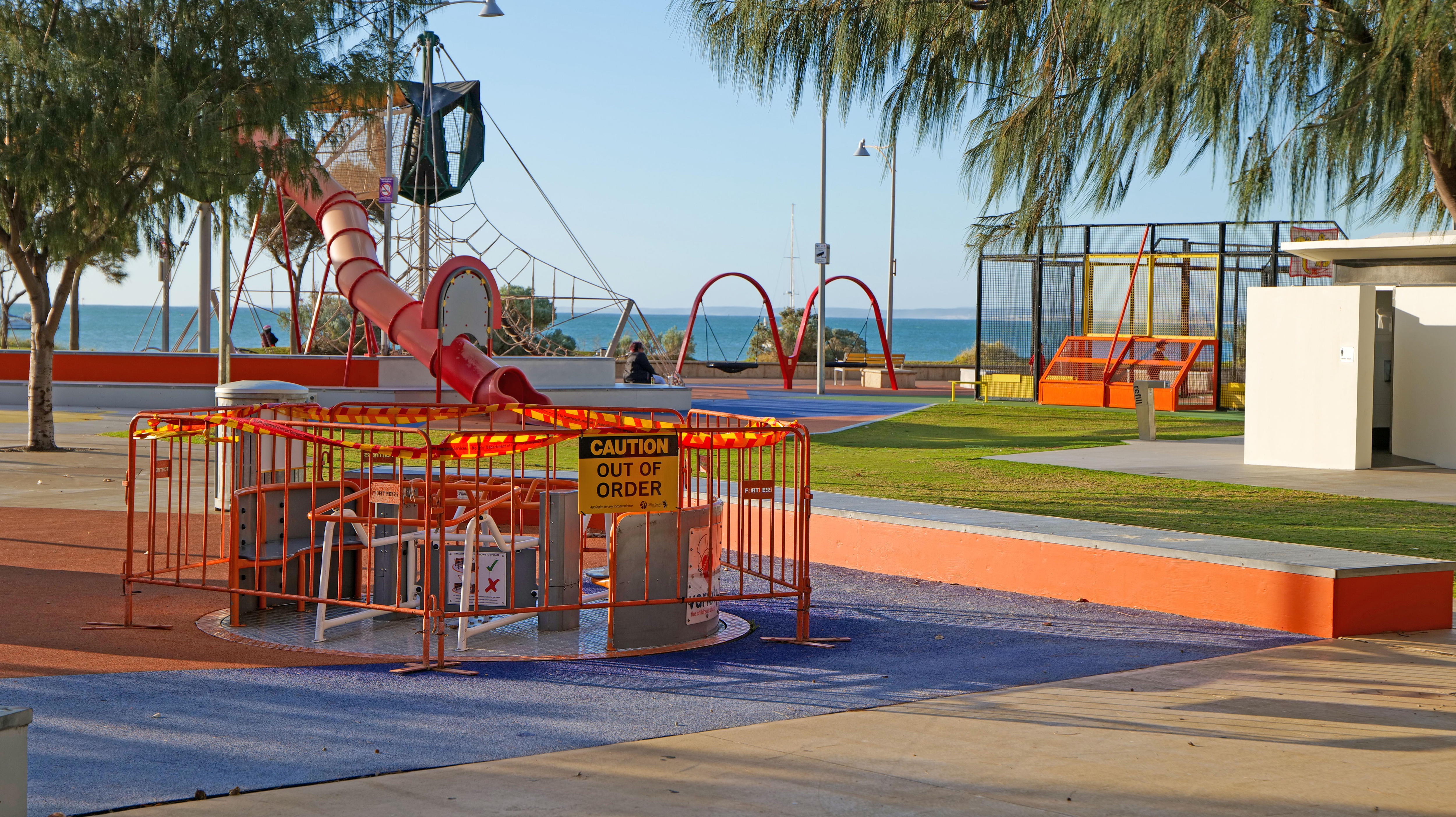 A colourful children's playground with ocean in background. A merry-go-round has barricades and caution tape 