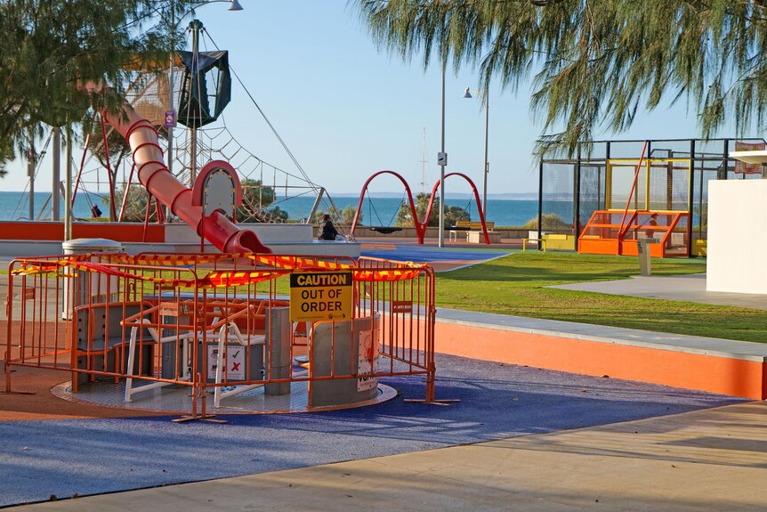 A colourful children's playground with ocean in background. A merry-go-round has barricades and caution tape 