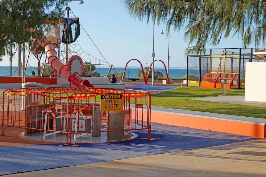 A colourful children's playground with ocean in background. A merry-go-round has barricades and caution tape 