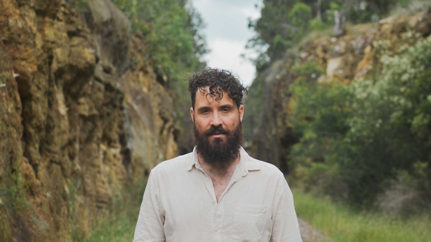 White man with curly dark brown hair and a full beard wearing a cream shirt and standing between two rocky cliffs