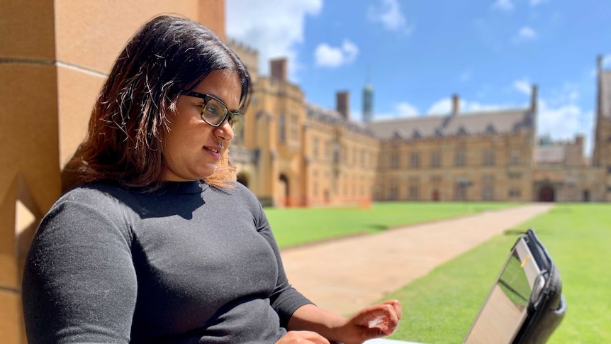 A young woman of Indian descent sits outside against a wall at a university studying on her laptop.