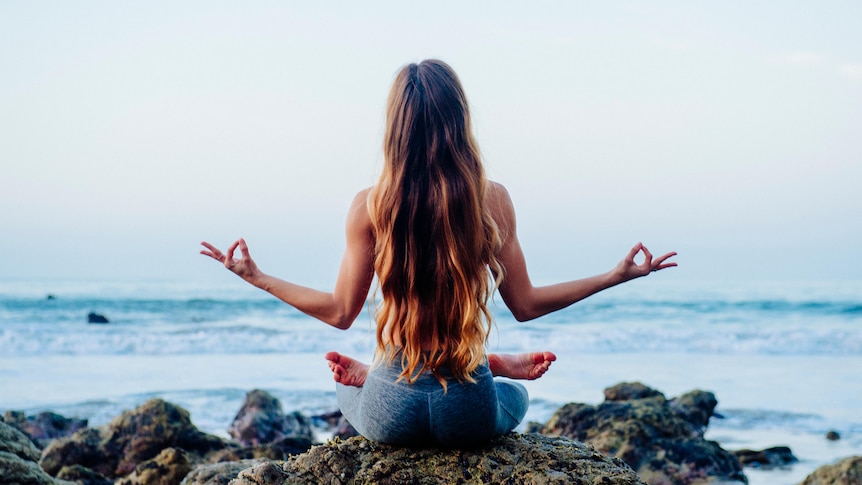 back of young woman doing yoga lotus pose on rock facing ocean waves