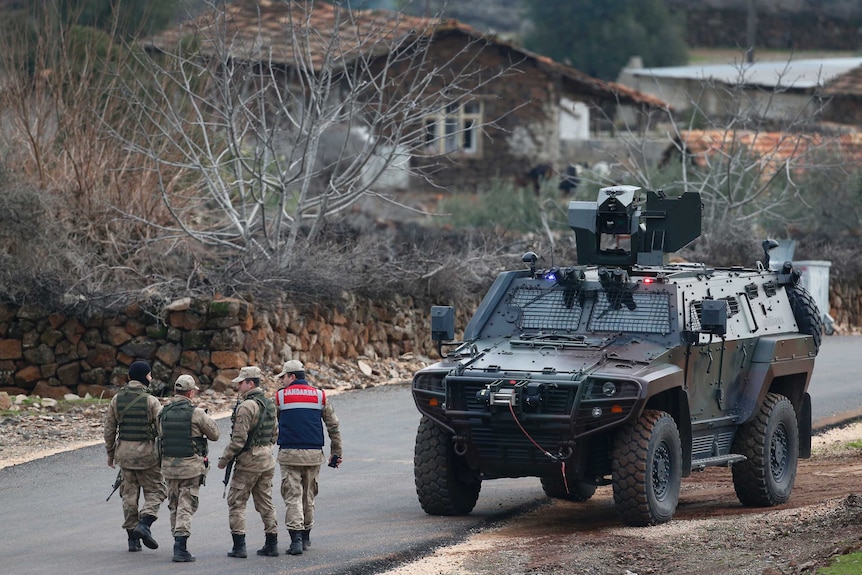 Wide shot of four men in army uniform walking near an armoured vehicle.