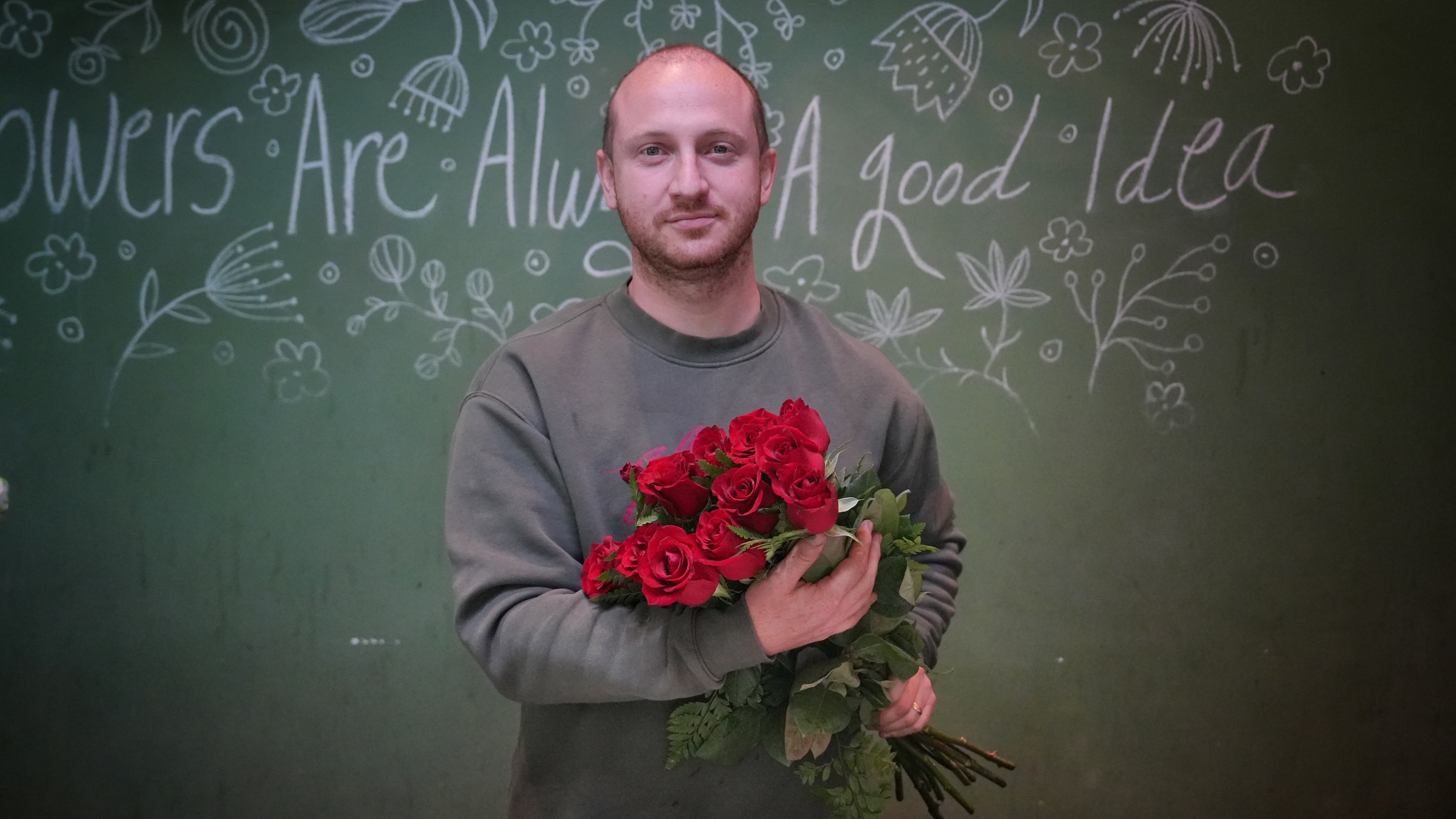 Man in grey jumper holding large bouquet of red roses with writing 'Flowers are always a good idea' in background