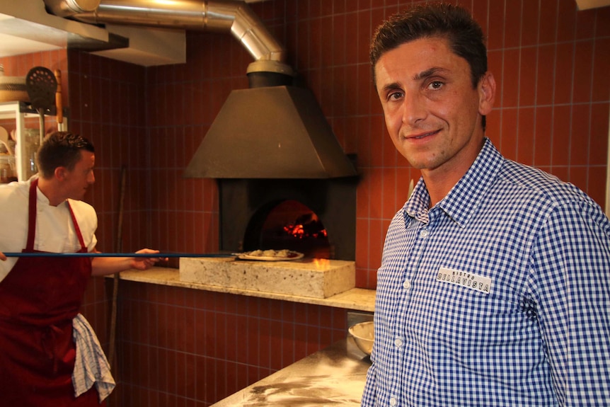 A man in a checked shirt stands next to a worker in an apron putting a pizza into a woodfired oven.