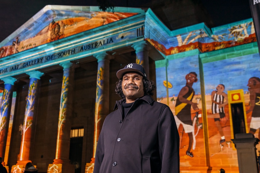 Vincent Namatjira out the front of the Art Gallery of SA at night, with a large projection of his work on display behind him