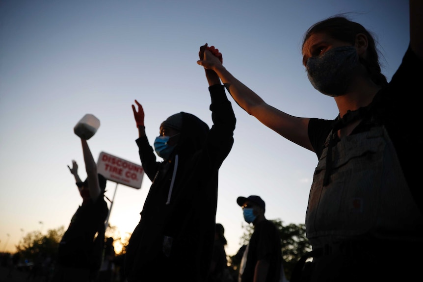 Three protesters are seen holding their hands up high, one holds a bottle with milk in it. The sky above indicates night's near.