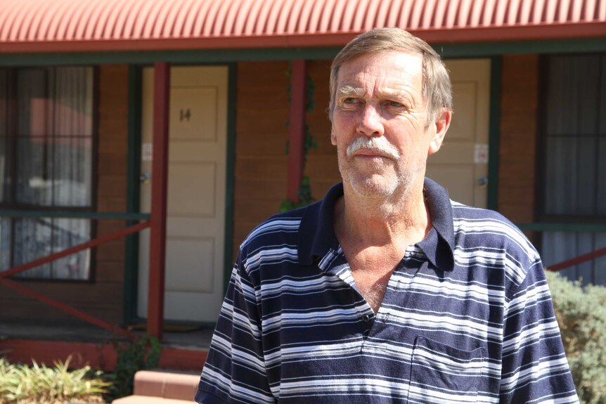 Man stands in front of a row of motel rooms in the car park of his motel, looking off-camera.