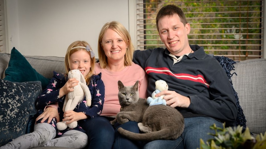 A woman with shoulder-length blonde hair sits on a couch beside a young blonde girl and a man. A grey cat sits on their lap.