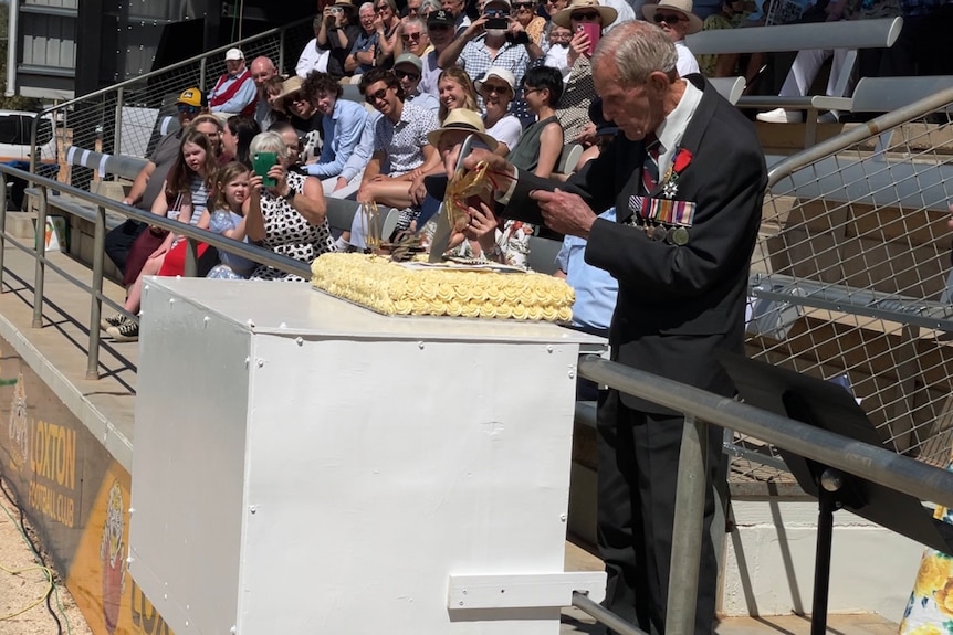 A elderly man, Howard Hendrick, with war medals on his suit cuts into a big yellow birthday cake in front of crowded bleachers.