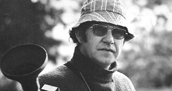 A man wearing a bucket hat and sunglasses stands in front of a leafy background.