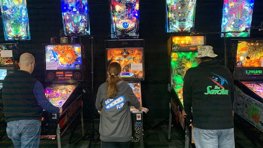 Three people from behind playing a pinball machine, the machines are very colourful.