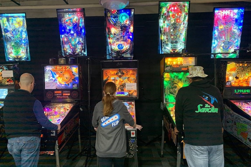 Three people from behind playing a pinball machine, the machines are very colourful.