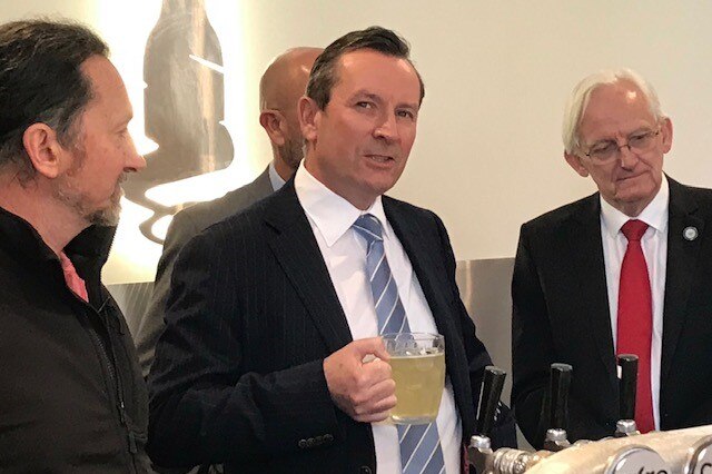 WA Premier Mark McGowan stands holding a mug of cider next to bar taps with three men surrounding him.
