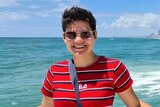 A woman in a red shirt and sunglasses smiles in front of the sea.