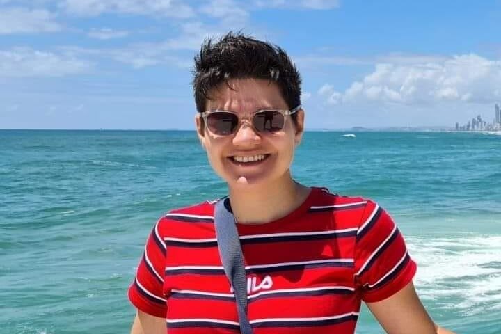 A woman in a red shirt and sunglasses smiles in front of the sea.