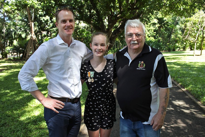 Two men and a young girl stand and smile at the camera. The girl is wearing war medals.