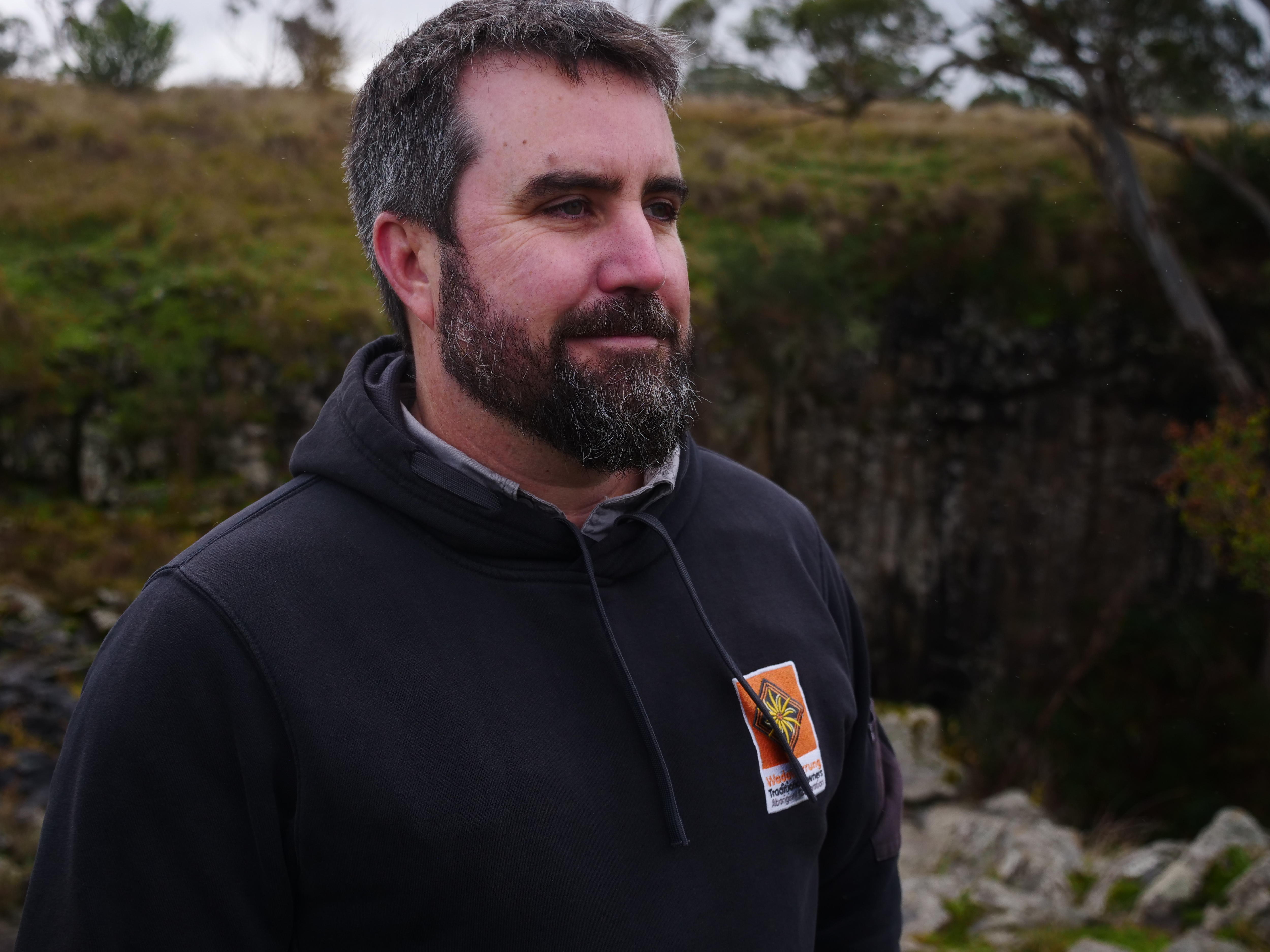 A man with a beard looks into the distance with greenery behind him