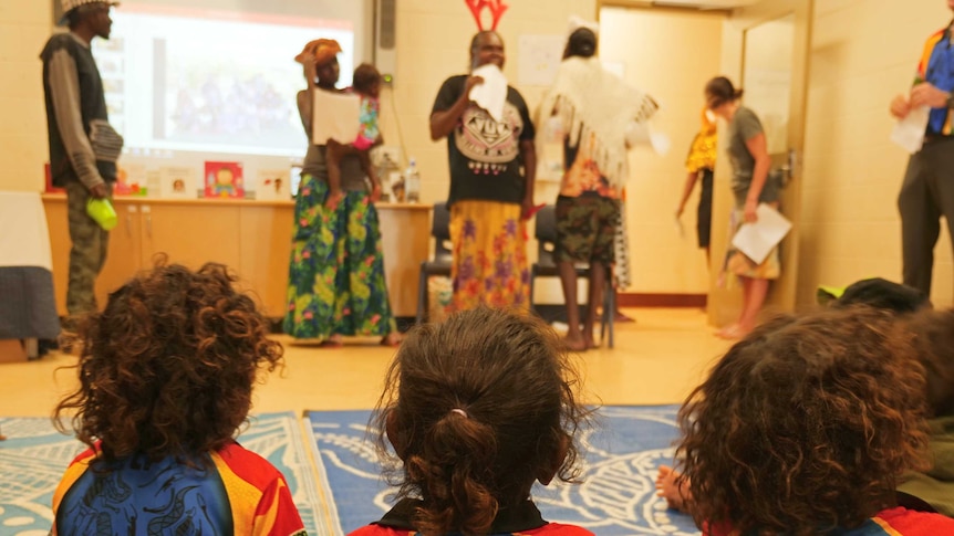 Three aboriginal children watching parents in homemade costumes perform in classroom.