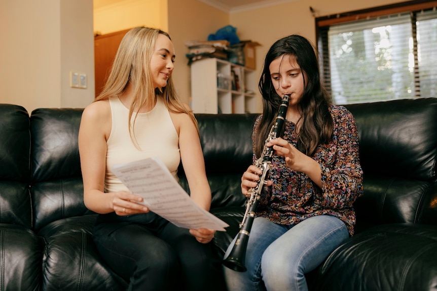 Woman sitting on a black leather couch holding a music manuscript, helping a young girl practise clarinet