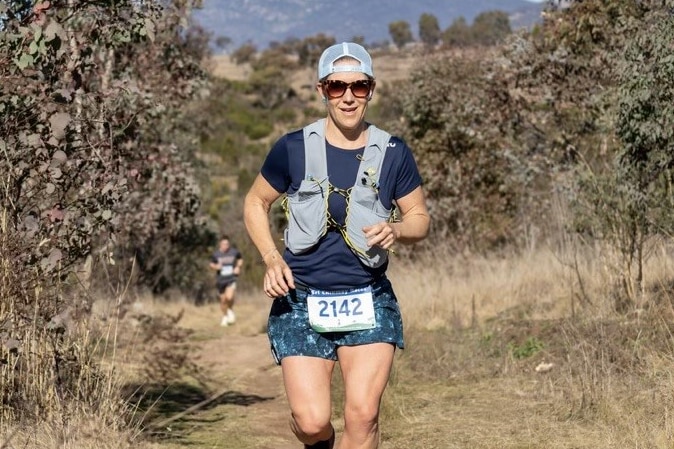A woman runs in the bush, wearing a drinks vest, sunglasses and a numbered bib, indicating she is competing in a race.