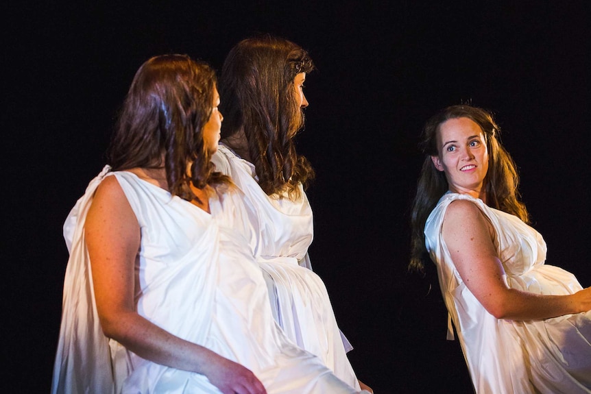 Three women in white dresses on stage, two look over at one who smiles back at them