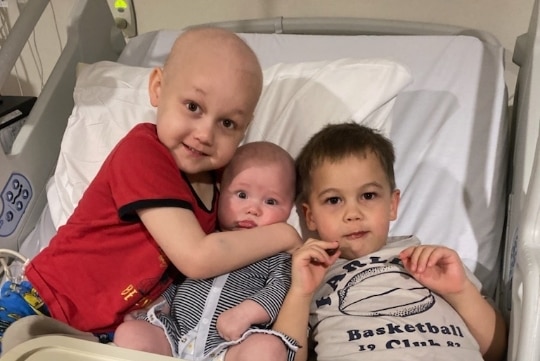 Three young children lay together on a hospital bed behind an M&M cake