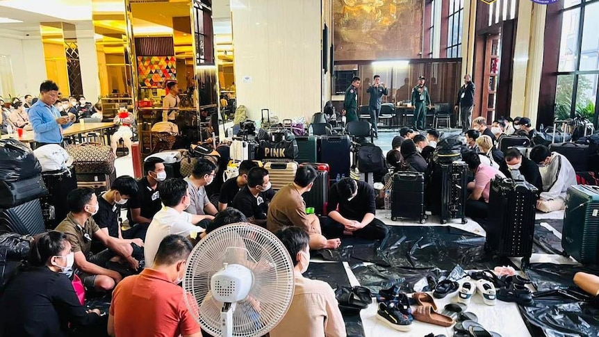 A large number of men sit on the floor of a hotel foyer around a pile of shoes with police officers stand in nearby.