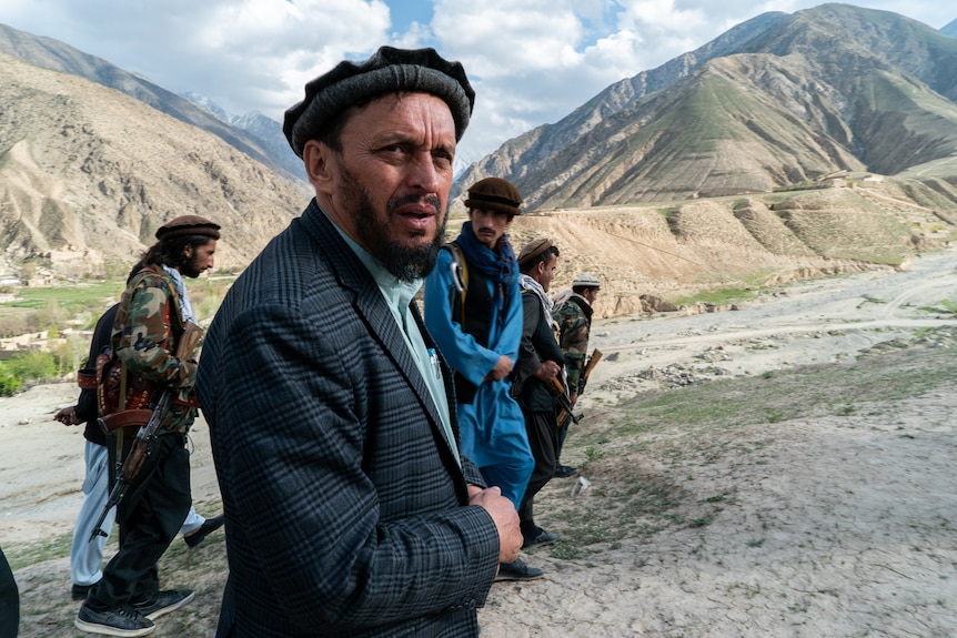 A man in a cap and jacket stands outside with mountains in the background