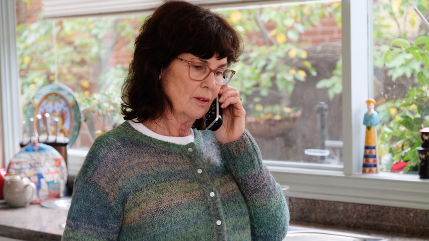 Jenny, dressed in a green and blue knitted top, stands in the kitchen and holds a mobile phone to her ear.
