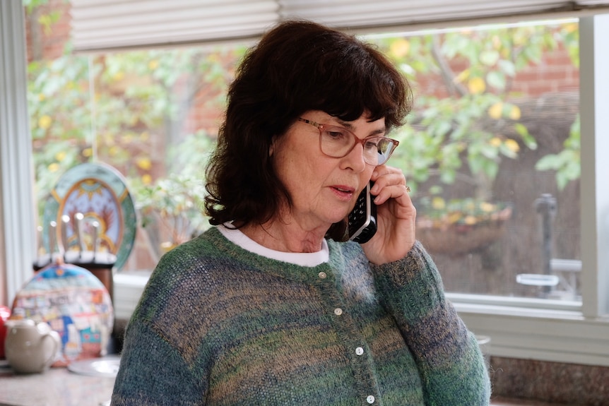 Jenny, dressed in a green and blue knitted top, stands in the kitchen and holds a mobile phone to her ear.