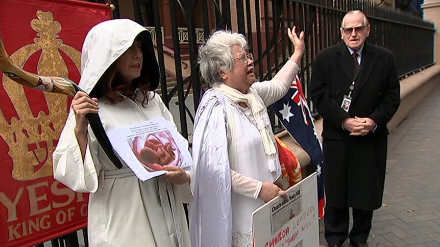 two women in white holding anti-abortion signs with older man in black coat and sunglasses next to them