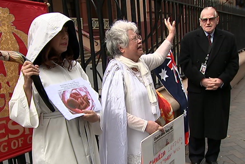 two women in white holding anti-abortion signs with older man in black coat and sunglasses next to them