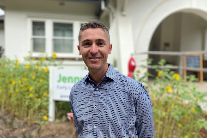 A guy looking at the camera smiling, standing outside a white building, being an Early Learning Centre