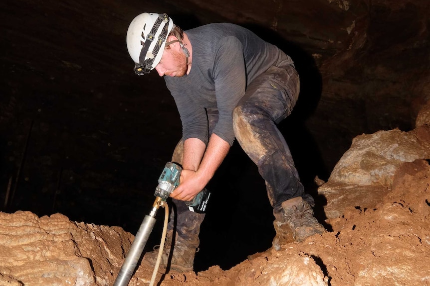 A bearded man in muddy clothes, boots and helmet operates a large drill inside a cave
