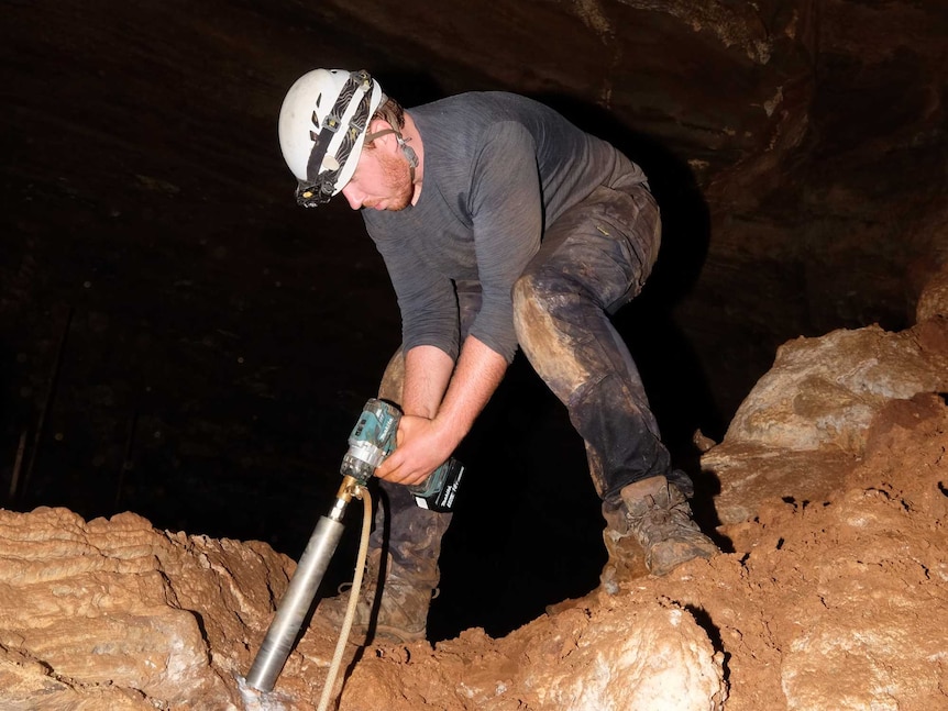 A bearded man in muddy clothes, boots and helmet operates a large drill inside a cave