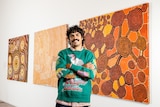 Presenter Tony Armstrong in a colourful jumper stands in front of a series of three First Nations artworks on a gallery wall.