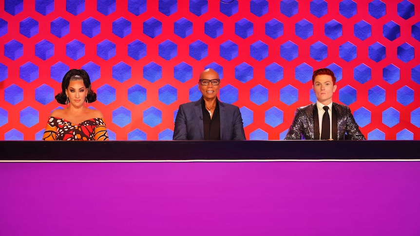 Michelle Visage (left), RuPaul Charles (centre) and Rhy Nicholson (right) sitting behind a desk.