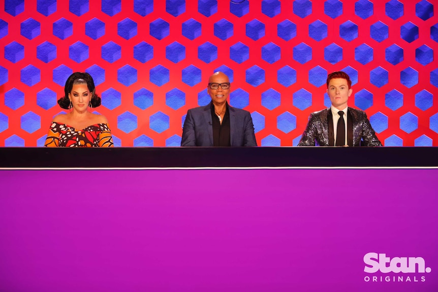 Michelle Visage (left), RuPaul Charles (centre) and Rhy Nicholson (right) sitting behind a desk.