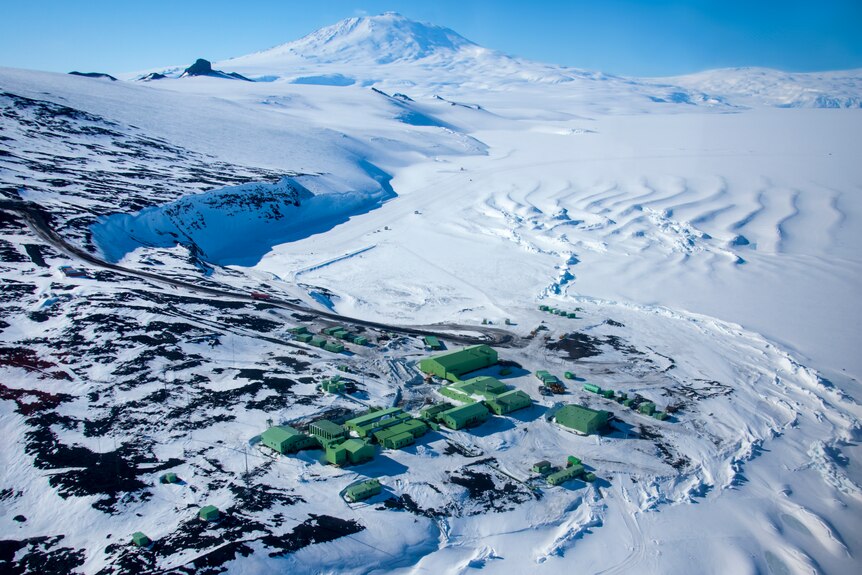 A set of squat, green buildings sits on a rocky, snow-covered plain in Antarctica.